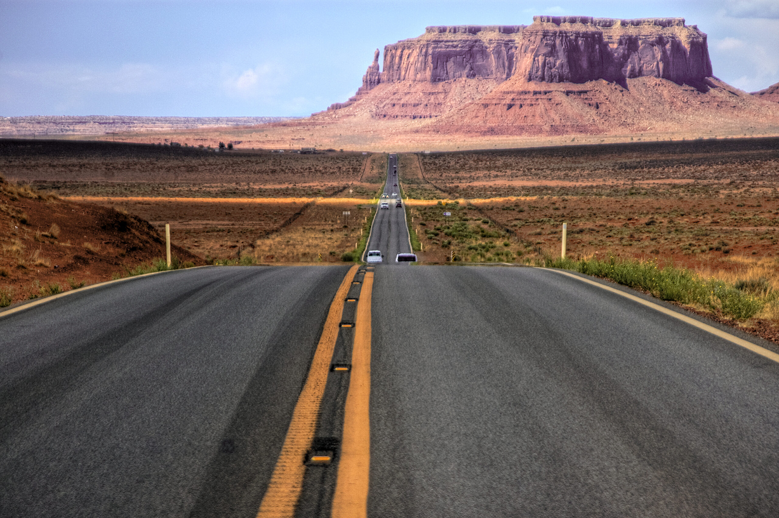 An empty stretch of U.S. Route 163 highway leading toward the red sandstone buttes of Monument Valley on the Utah–Arizona border, an iconic American road-trip landscape.
