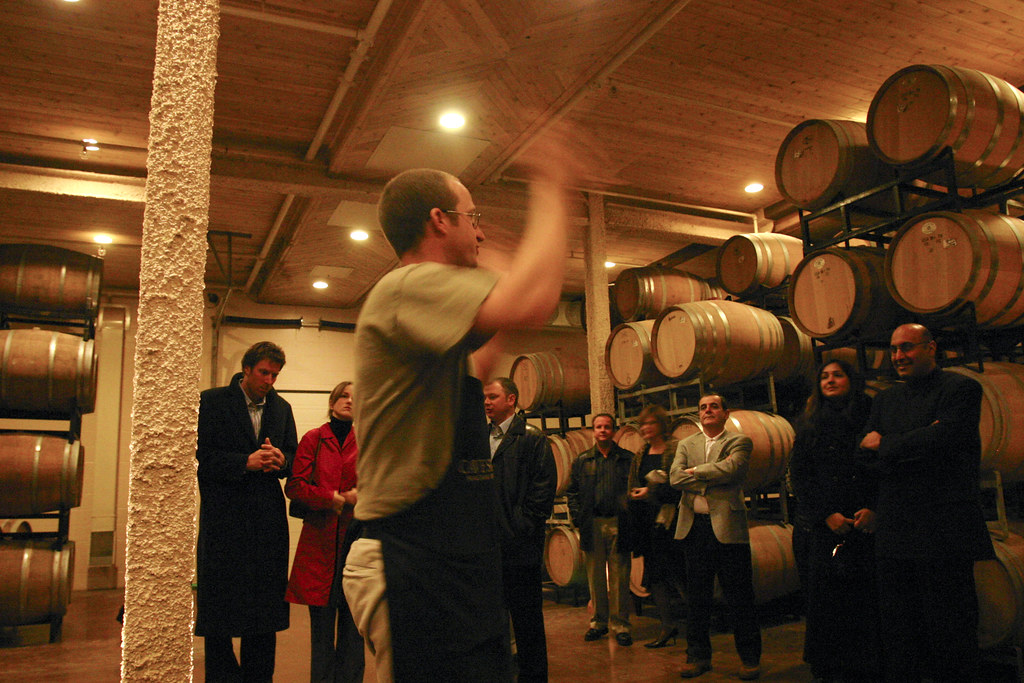 Rows of oak wine-ageing barrels in the cellar at Cave Spring winery in Jordan Village, Lincoln, Ontario, with a guide explaining the barrel-ageing process to visitors.