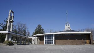St. George Roman Catholic Church in Crystal Beach, Ontario: a low mid-century-modern brick church with a curved white roofline, a tall metal globe-and-cross spire, and a tall freestanding concrete bell tower to the left, under a clear blue sky.