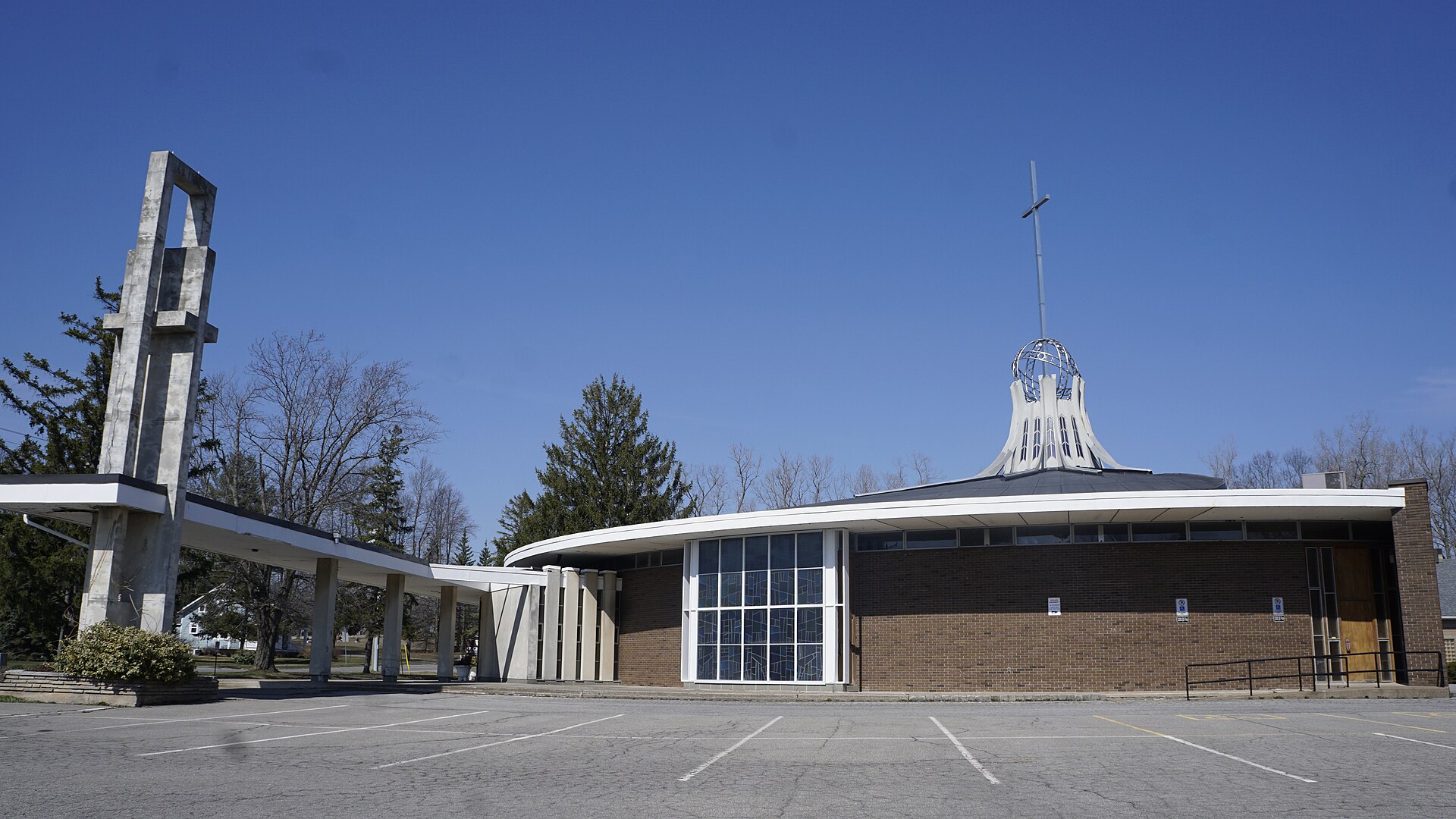 St. George Roman Catholic Church in Crystal Beach, Ontario: a low mid-century-modern brick church with a curved white roofline, a tall metal globe-and-cross spire, and a tall freestanding concrete bell tower to the left, under a clear blue sky.