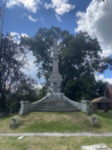The grey granite Lundy's Lane Battlefield Soldiers' Monument, a tall obelisk on a wide stepped base inscribed "LUNDY'S LANE" and "1812-14", flanked by cannonballs with a Union Jack flag at left and a red-brick chapel at right, set among trees at Drummond Hill Cemetery in Niagara Falls, Ontario.