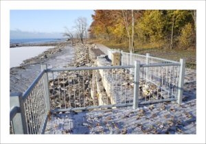 Paved lakeside path with a galvanized metal railing running along the rocky north shore of Lake Erie on the Friendship Trail at Waverly Beach in Fort Erie, Ontario, framed by autumn-colored trees on a clear day.