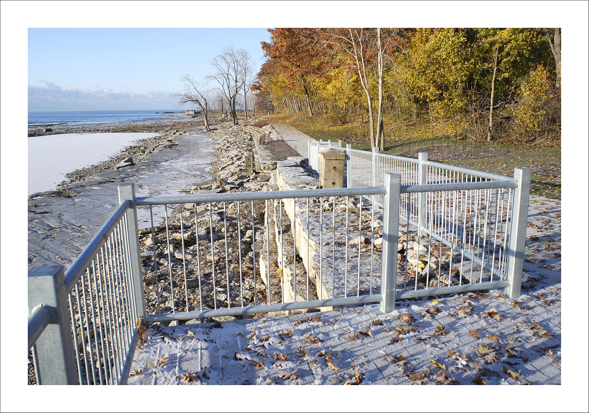Paved lakeside path with a galvanized metal railing running along the rocky north shore of Lake Erie on the Friendship Trail at Waverly Beach in Fort Erie, Ontario, framed by autumn-colored trees on a clear day.