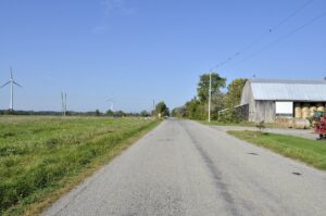 A quiet rural road running through flat West Lincoln farmland, with a weathered wooden barn, round hay bales, and distant wind turbines under a clear sky — typical countryside of the Niagara-region township that surrounds the hamlet of St. Anns.
