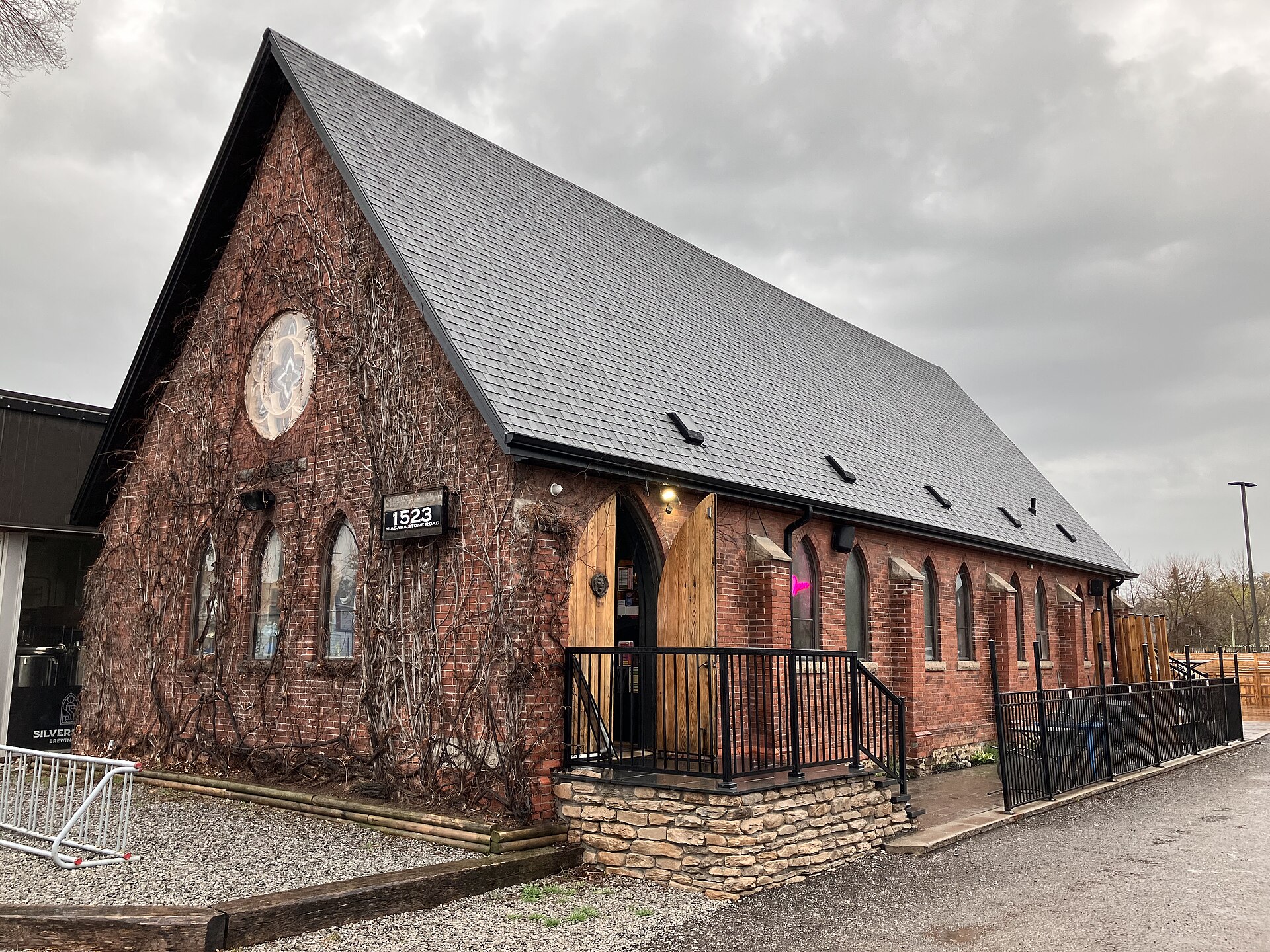 The red-brick, Gothic-Revival former Anglican church that houses Silversmith Brewing Co. in Virgil, Ontario, with pointed-arch windows, an ivy-covered gable, a rose-window medallion, and a black sign reading "1523 Niagara Stone Road" beside its wooden arched doorway.