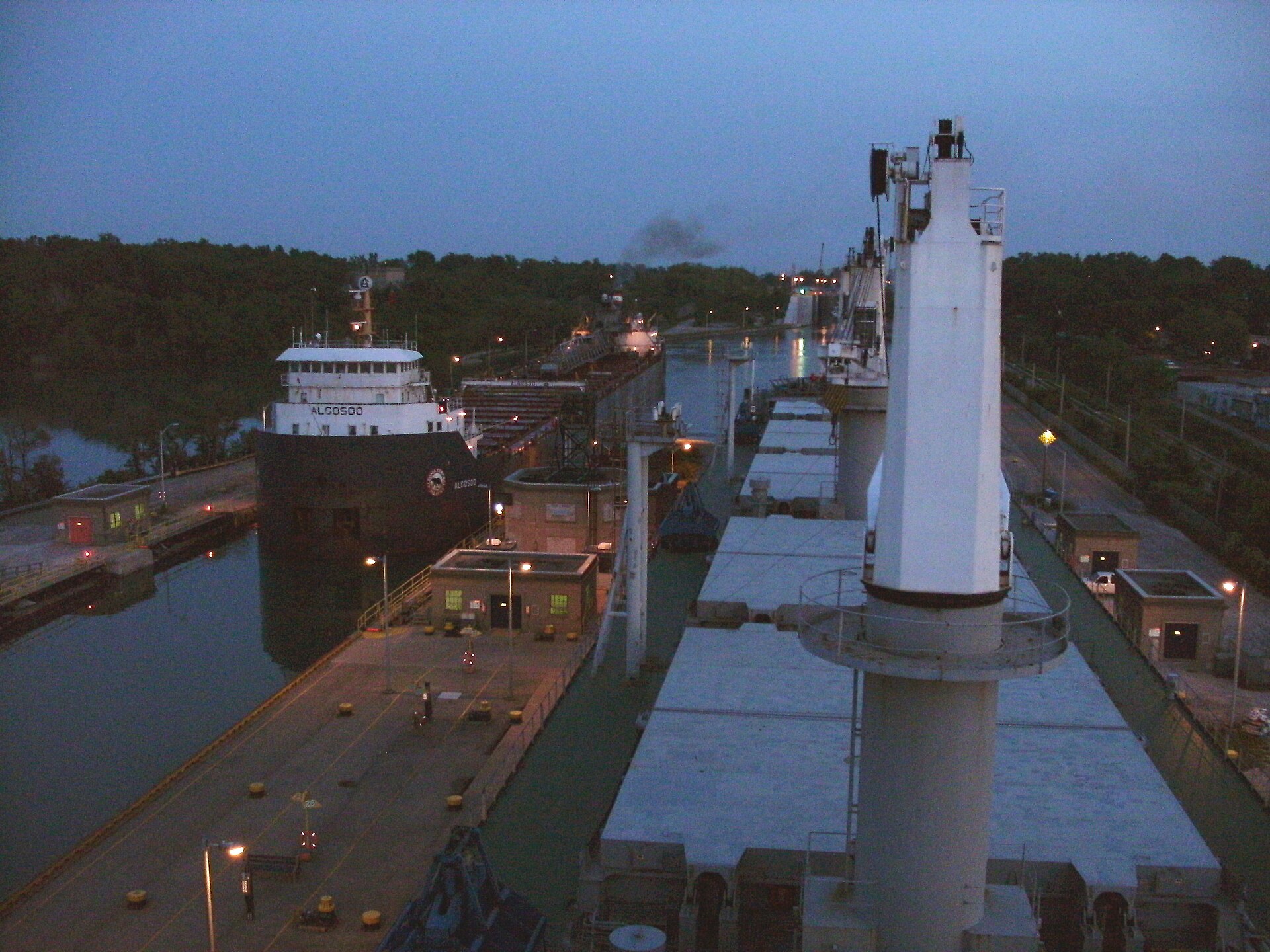 Two Great Lakes freighters pass each other in the parallel chambers of the Welland Canal's twin flight locks at Thorold, Ontario, at dusk, with lockside control buildings and the canal beyond.